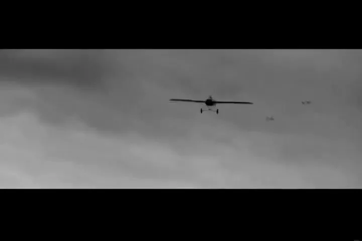 A pilot in a biplane flies through a cloudy sky, with other planes visible in the distance, in a black and white shot.