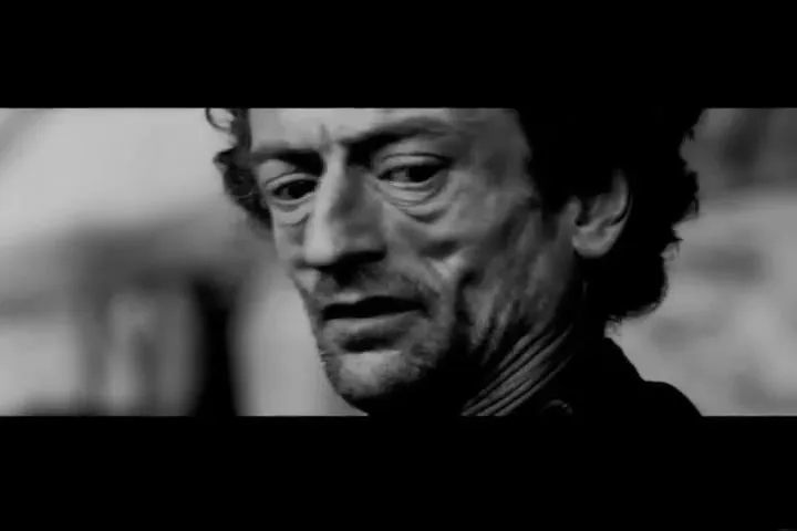 A gaunt, middle-aged man with curly hair and a beard looks around nervously in a black and white, dimly lit interior.