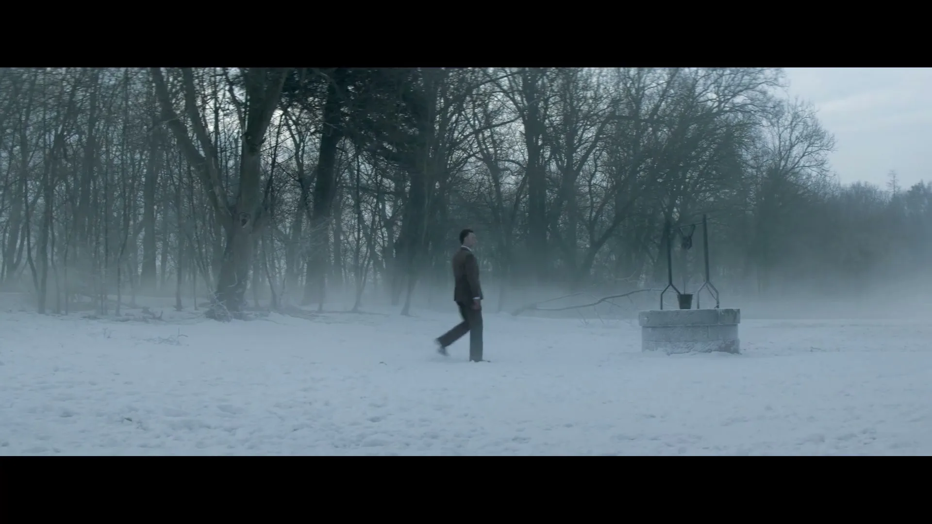 A man in a brown suit walks across a snowy field towards a well, with a misty forest in the background on a cold day.
