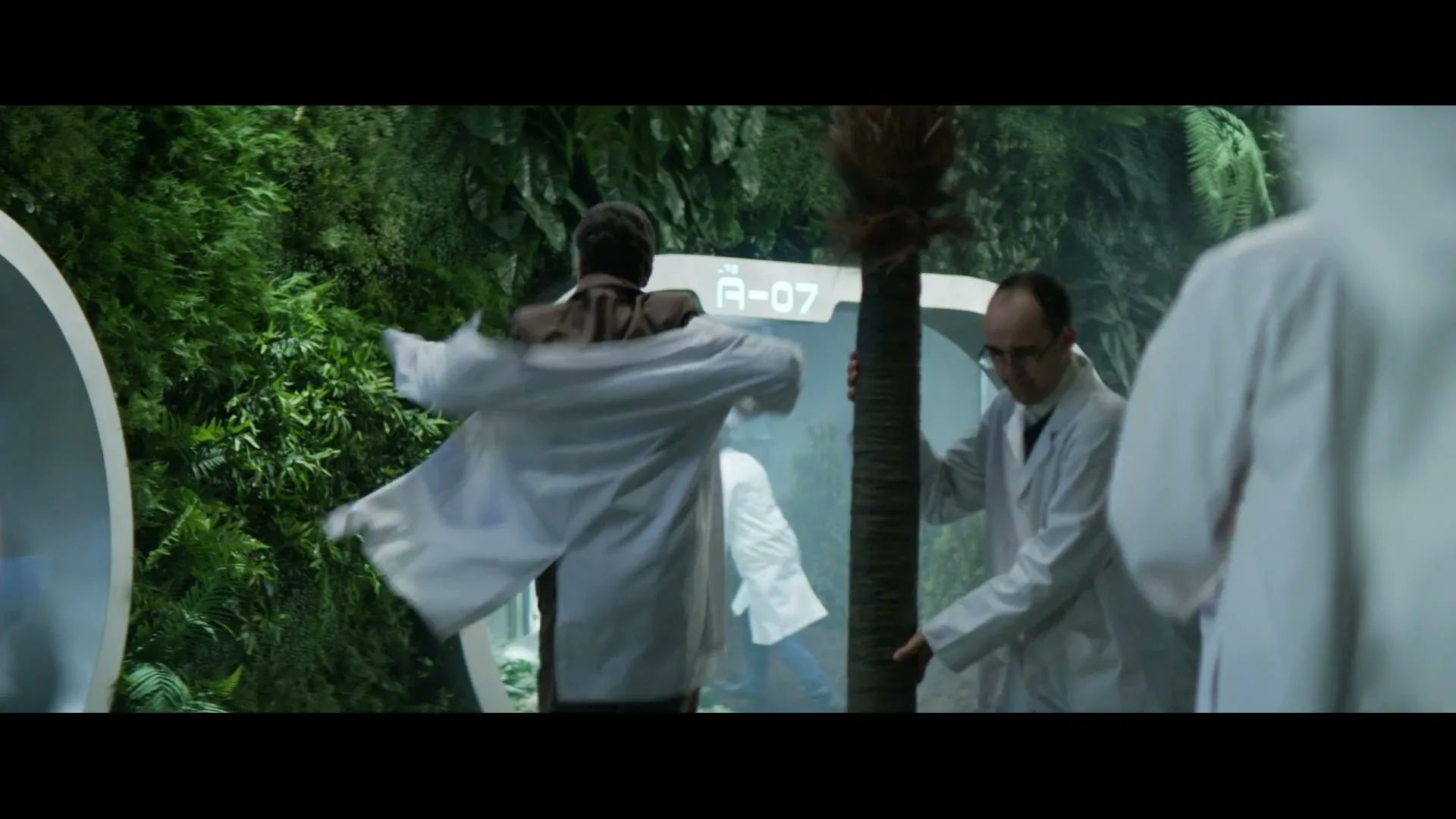 A man in a brown shirt puts on a white lab coat while other scientists in lab coats move a small palm tree in a futuristic lab with green walls.
