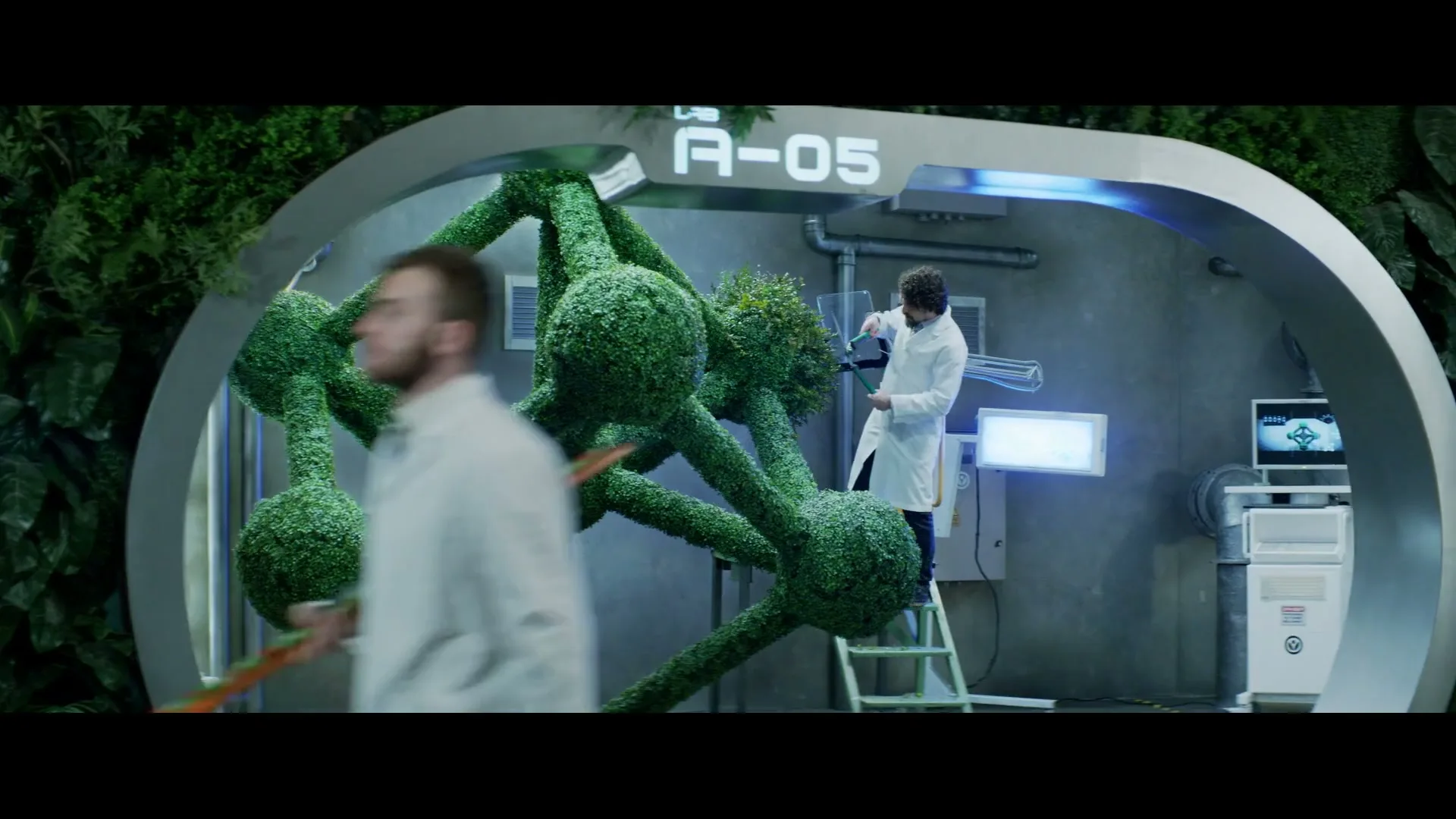 Two male scientists in lab coats work on a large, green, molecular-shaped topiary in a futuristic lab with plants on the walls.