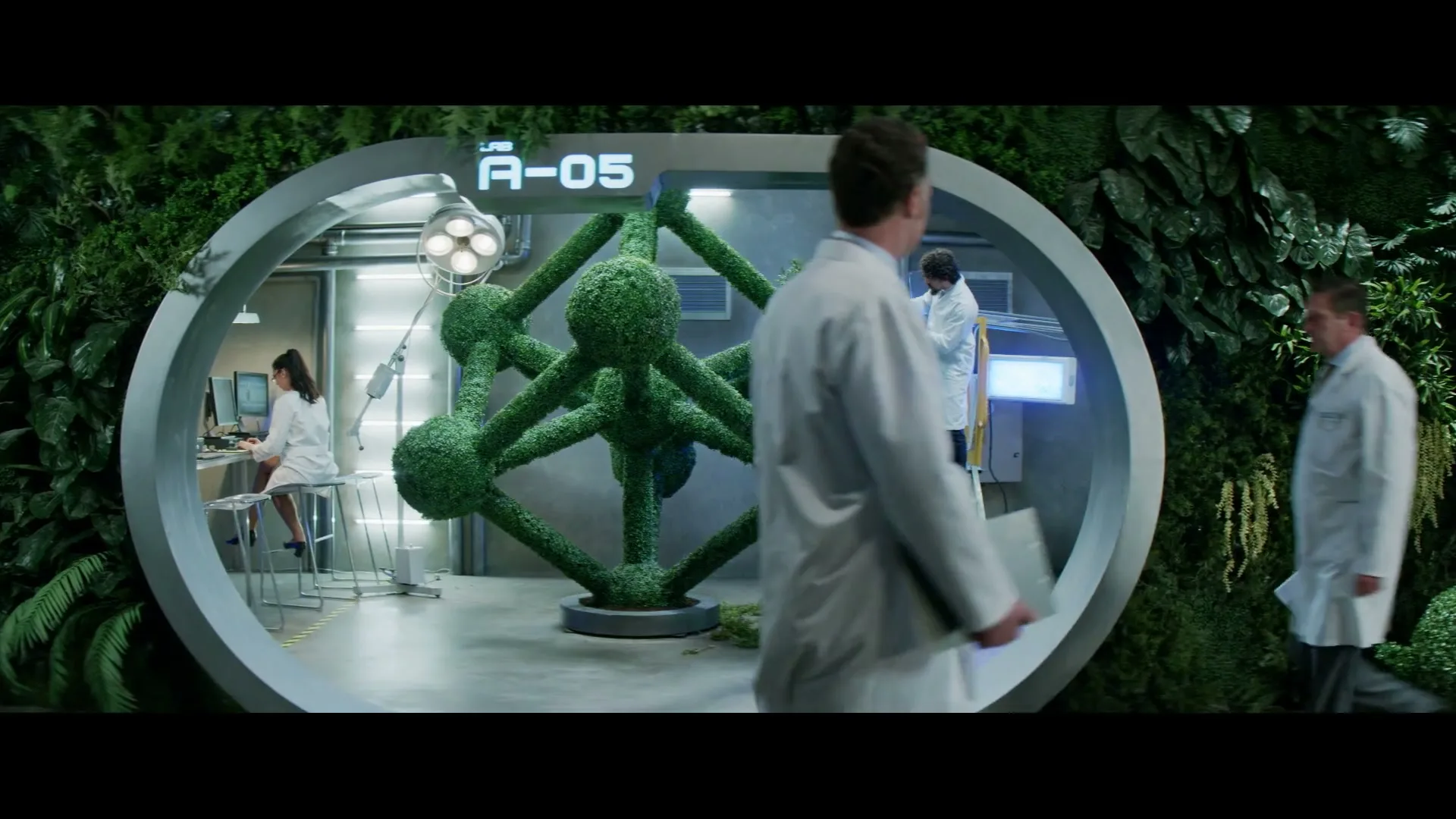 Scientists in lab coats work in a futuristic lab with a large topiary sculpture, surrounded by lush green walls.