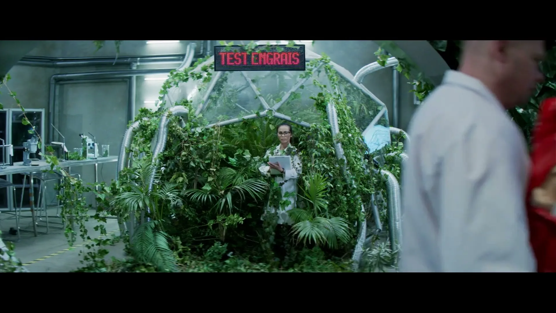 A male scientist in a lab coat walks past a female scientist standing inside a glass dome filled with plants, holding a tablet.