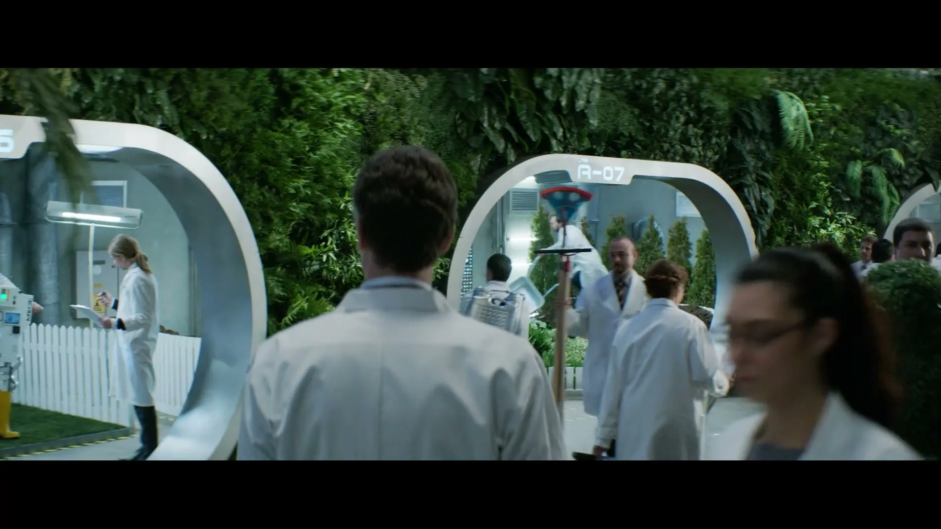A group of scientists in lab coats walk through a futuristic garden facility, some carrying gardening tools and plants, as one scientist walks away from the camera.