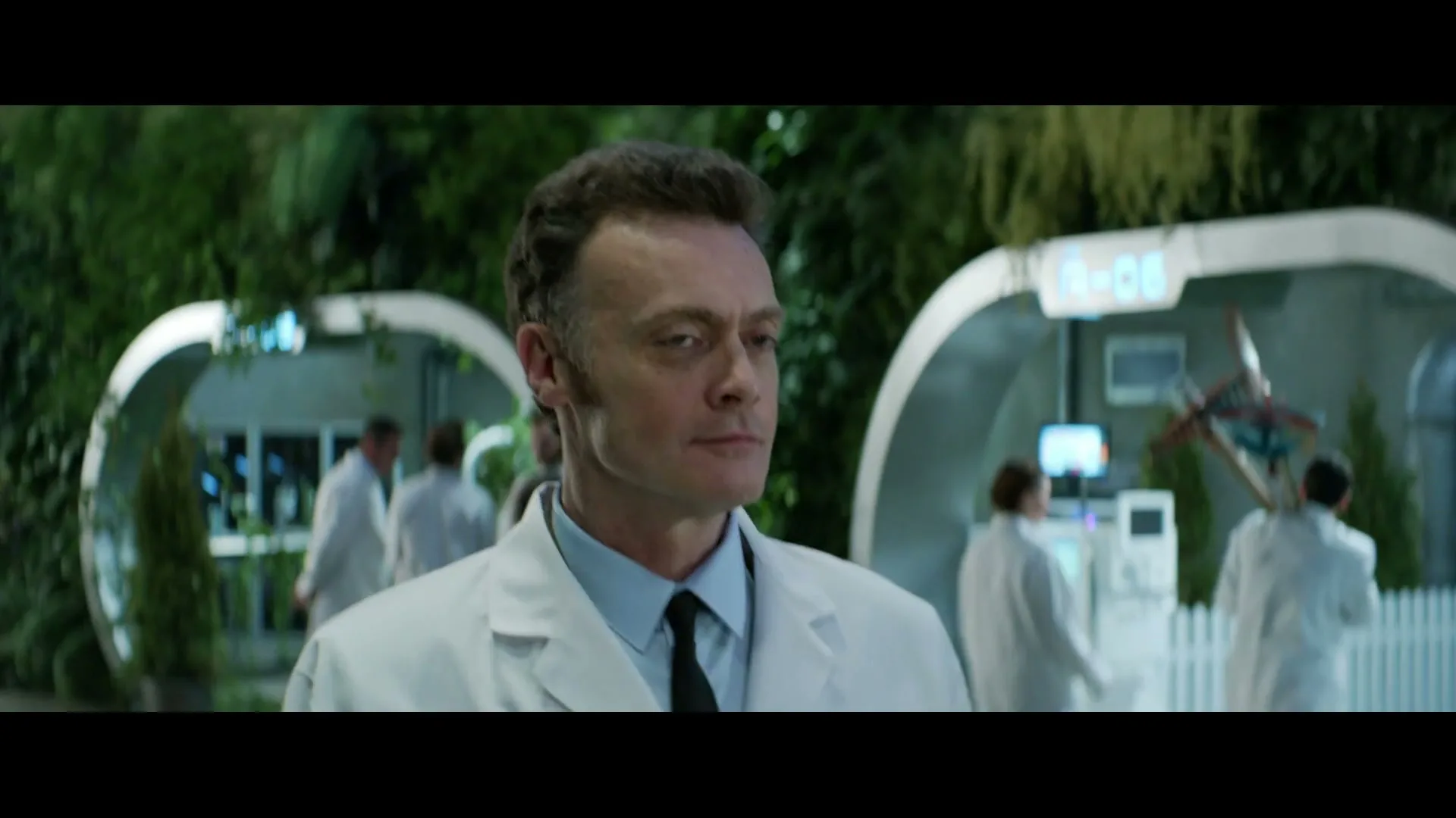 A male scientist in a lab coat looks around a futuristic, plant-filled laboratory with other scientists in the background.