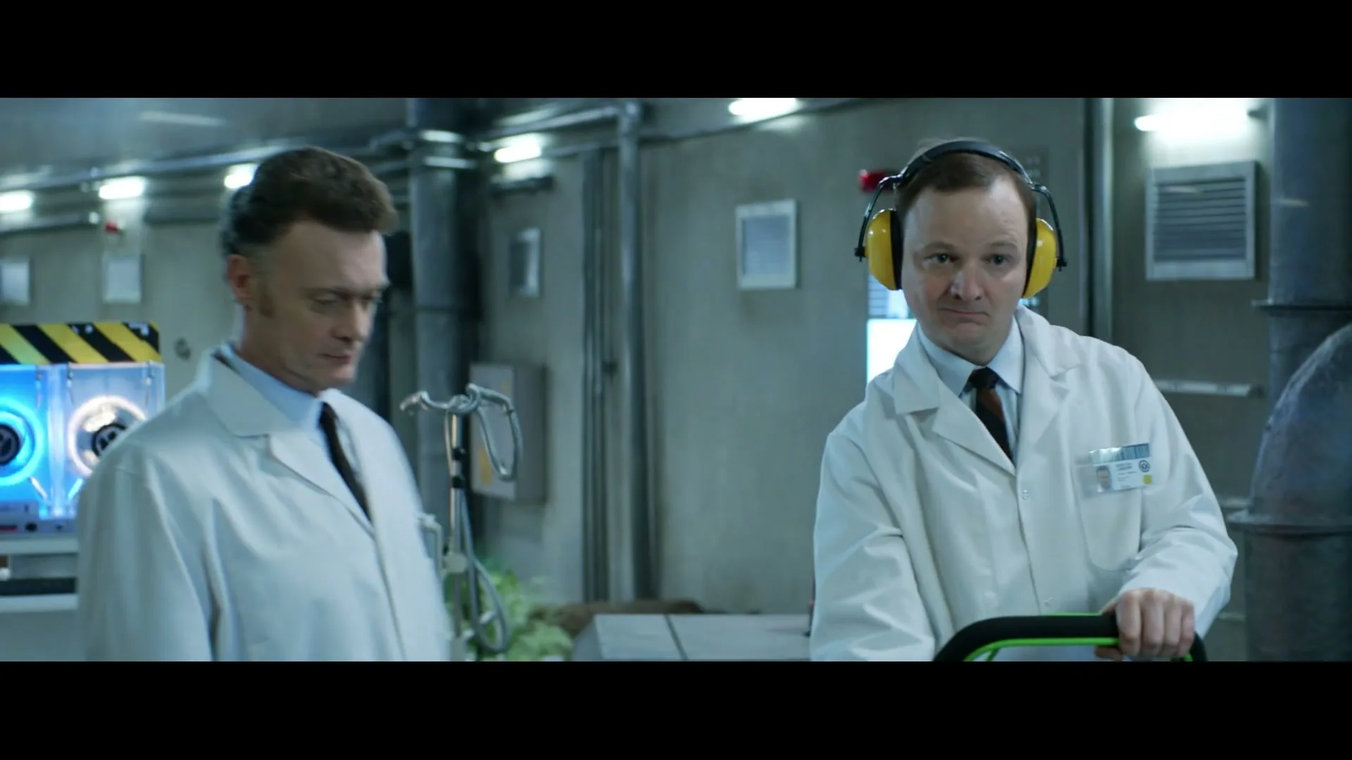 Two male scientists in lab coats and ear protection operate a machine in a futuristic lab, looking down at their work.