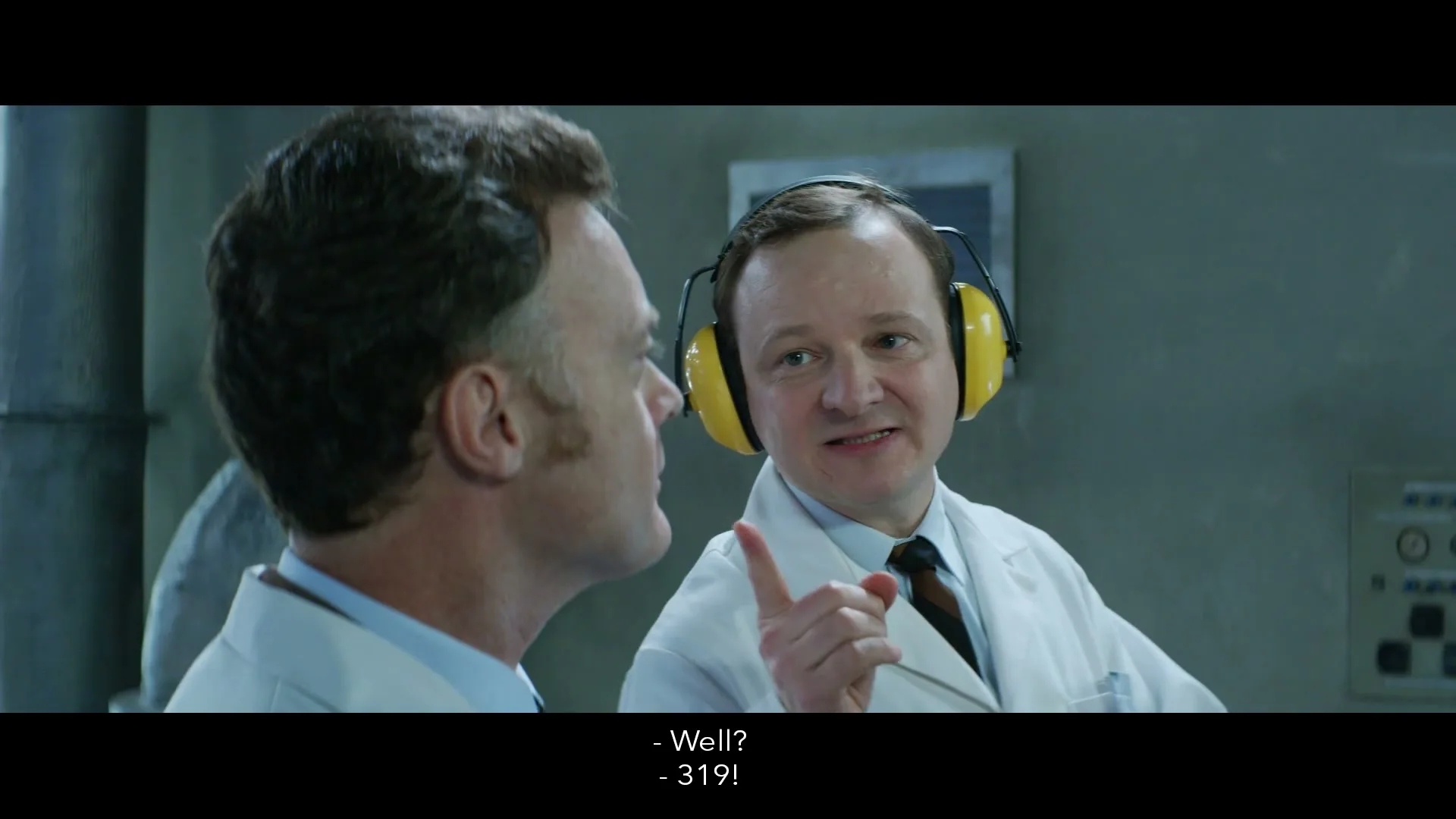 Two male scientists in lab coats and yellow ear protectors talk in a lab, one points his finger while speaking.