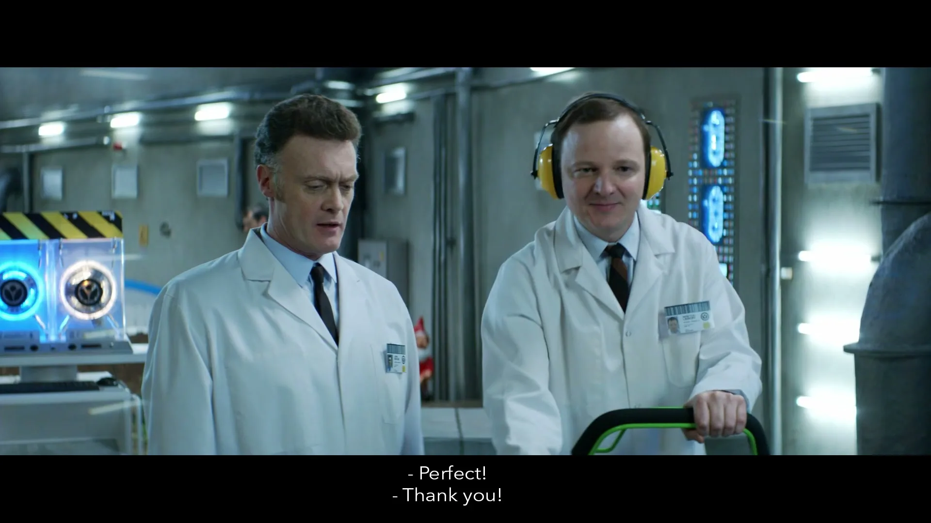 Two male scientists in lab coats and ear protection stand in a futuristic lab, one holding a green device, as they look at something off-screen.