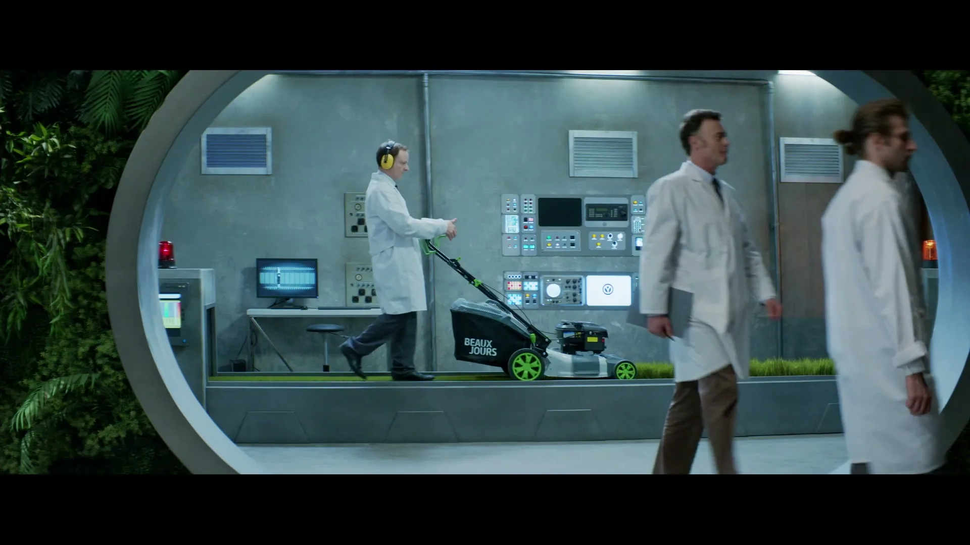 Three male scientists in lab coats are in a futuristic lab, with one pushing a lawnmower on a strip of grass, while two others walk past.