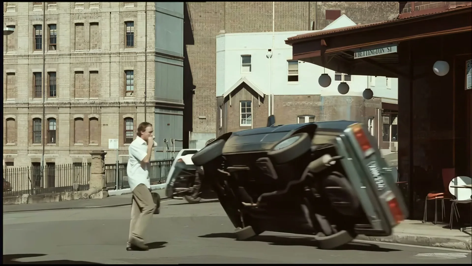 A man in a white shirt and khaki pants walks across a city street as cars flip and crash around him, while another man sets up a cafe table.