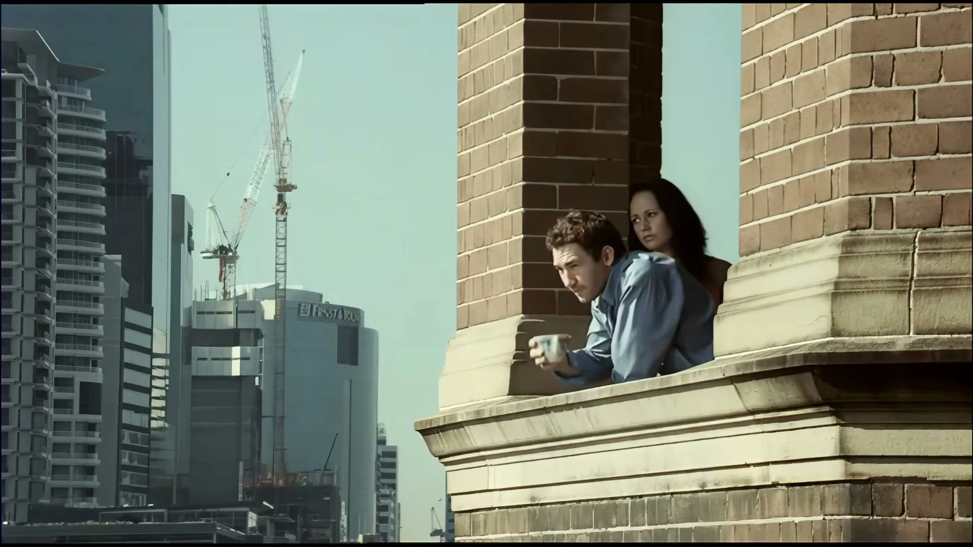 A man and a woman look out of a window in a brick building, observing a city skyline with skyscrapers and construction cranes in the background.