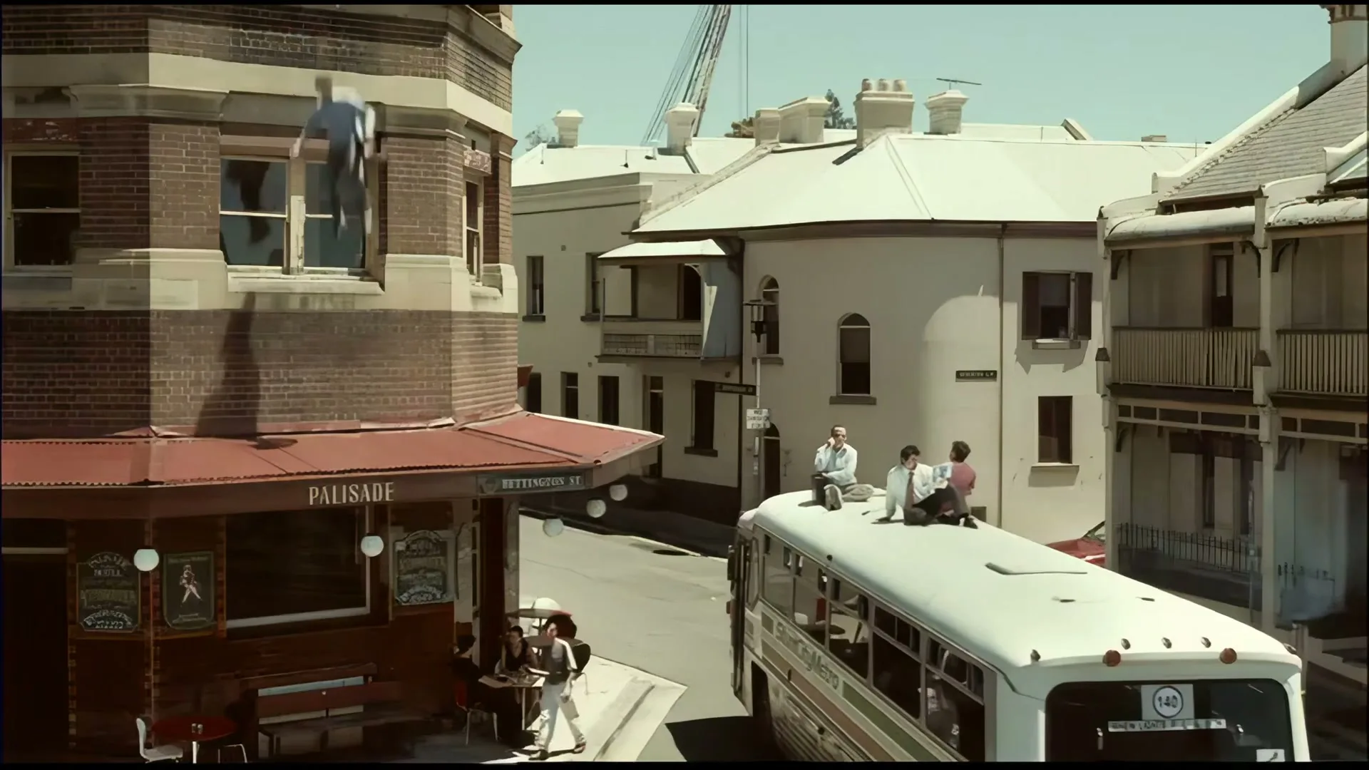 A man in a blue shirt hangs from a building ledge, while people sit on a bus roof and at a cafe table on a sunny street.