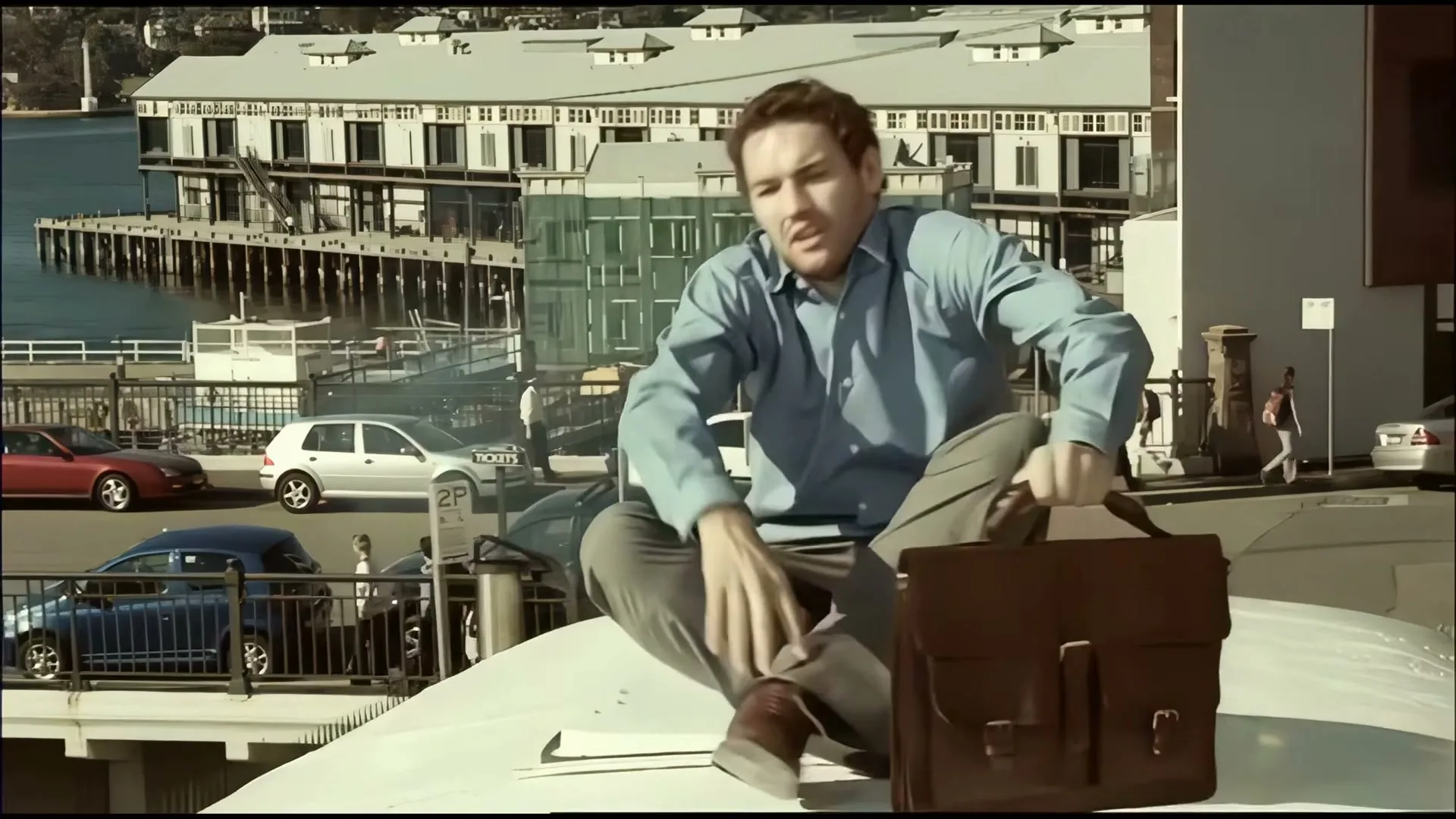 A man in a blue shirt and grey pants jumps onto the roof of a white van, holding a brown briefcase, with a harbor and buildings in the background.