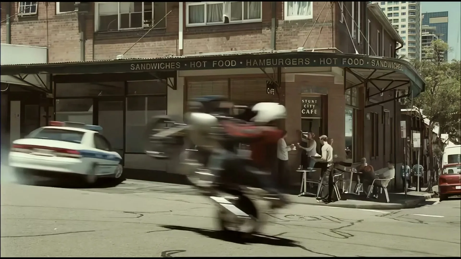 A police car drifts around a corner, kicking up smoke, as a motorcycle speeds past on one wheel, all in front of a busy cafe.
