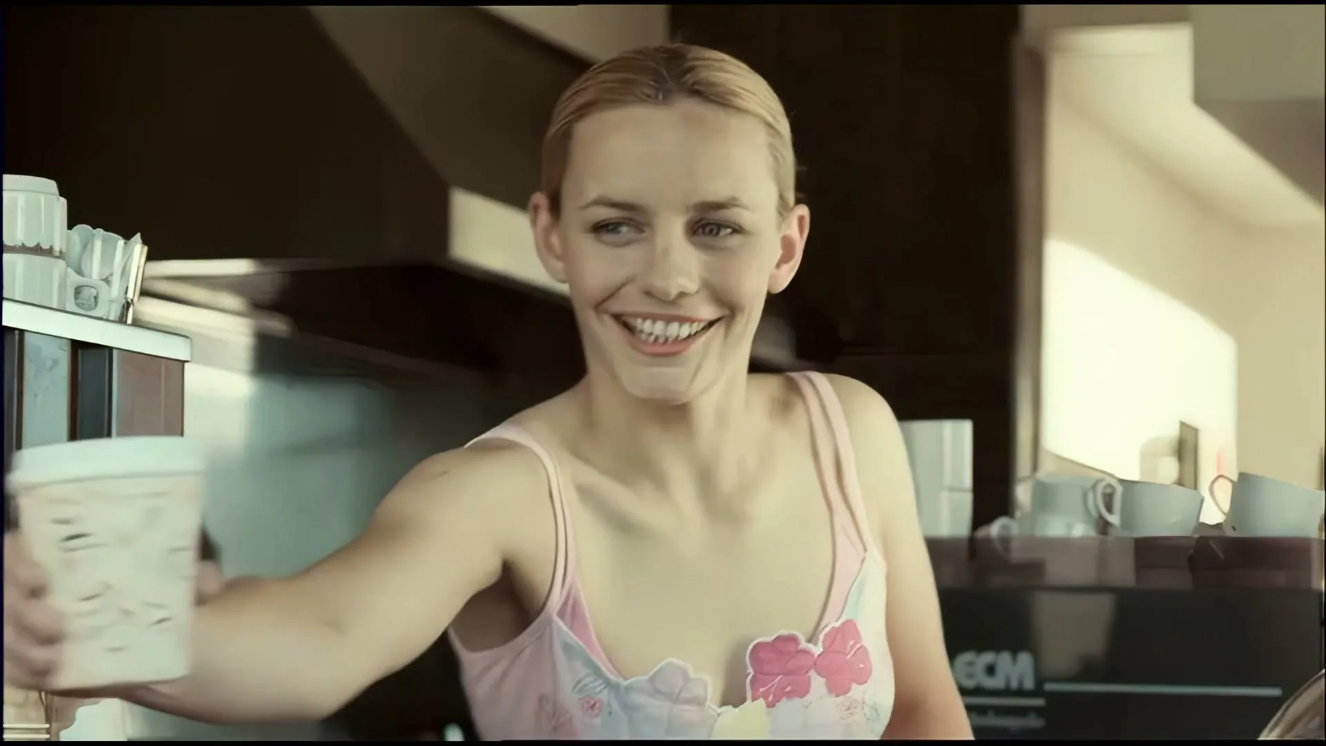A young woman with blonde hair, wearing a pink top, smiles while working behind a counter in a cafe.