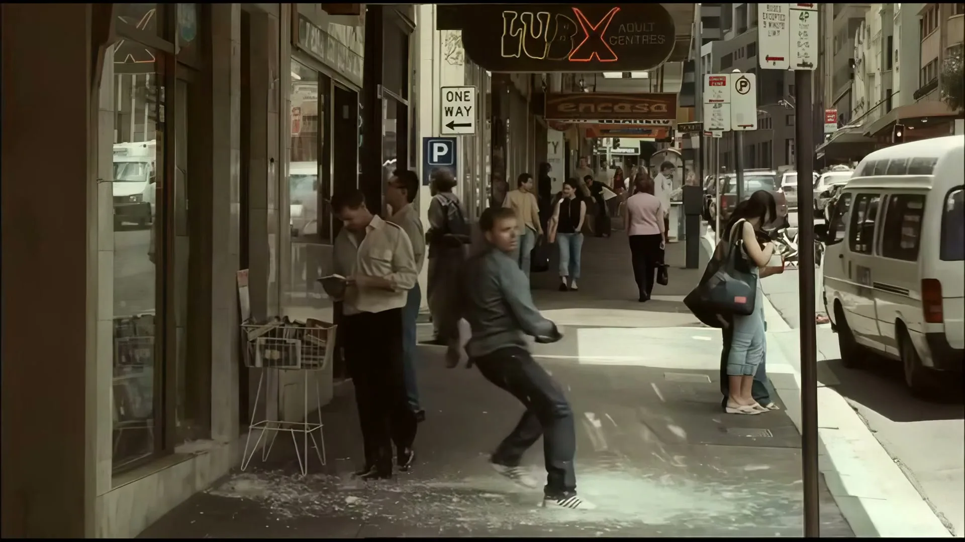 A man in a grey shirt and dark pants slips on a white substance on a busy city sidewalk, scattering items from a nearby display.