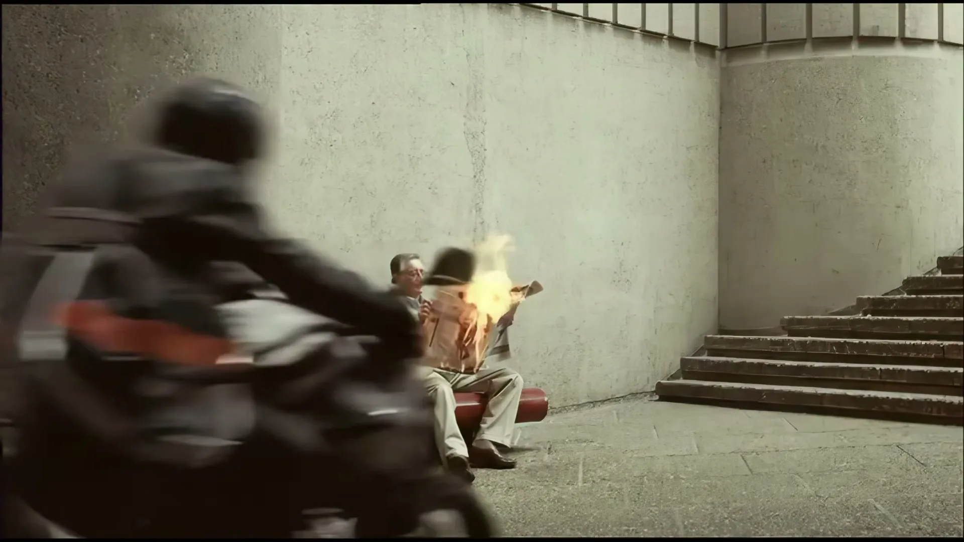 A man in a suit sits on a bench, reading a newspaper that is on fire, while a woman sits next to him and a motorcycle speeds past.