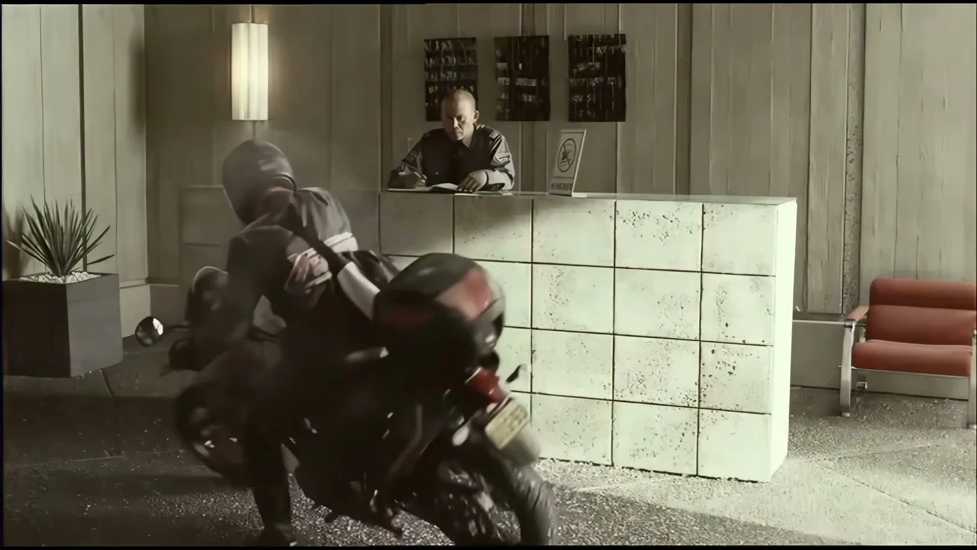 A motorcyclist in a helmet and black gear skids to a stop in front of a reception desk, where a man in a suit is writing.