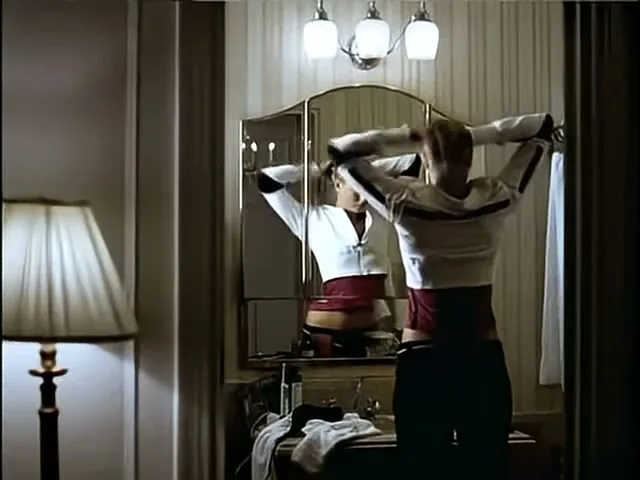 A young woman with blonde hair, wearing a white jacket and red top, stands with her back to the camera, looking at her reflection in a dressing table mirror in a dimly lit room.