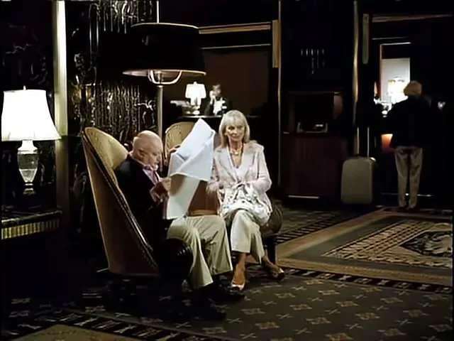 An older man reads a newspaper while an older woman sits beside him in a luxurious hotel lobby, with a blurred figure in the background.