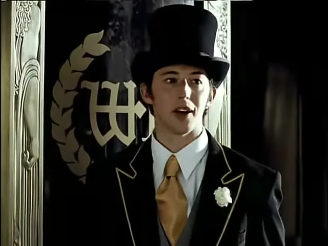 A young man in a top hat and formal wear stands in front of a dark background with a gold emblem, looking surprised.