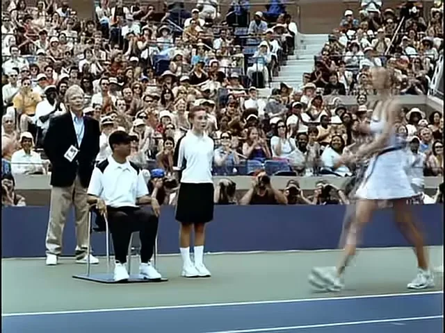A female tennis player in a white dress walks across a tennis court, past a man in a suit and two ball boys, with a crowd in the background.