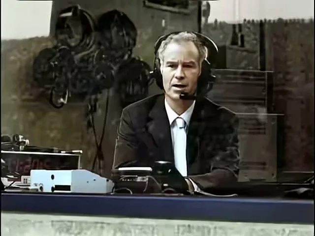 A male sports commentator in a suit and headset sits at a desk, speaking into a microphone in a broadcast booth.