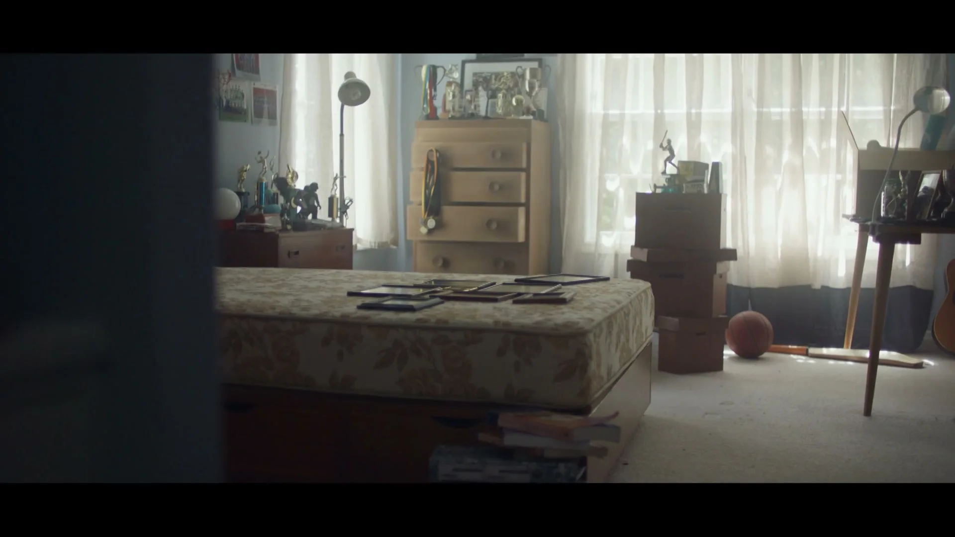 A wide shot of a messy teenage boy's bedroom with a bed, dresser, and desk, filled with trophies and sports equipment, seen from a slightly obscured doorway.
