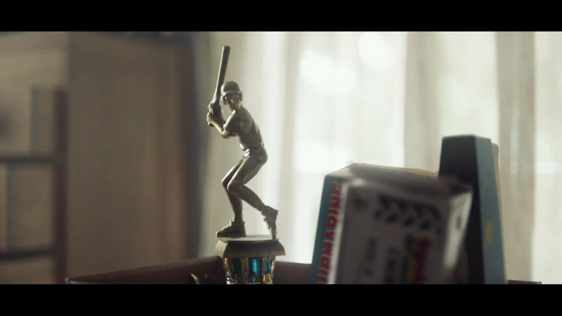 A baseball trophy stands on a shelf next to other items, with a window in the background, in a slightly dark room.