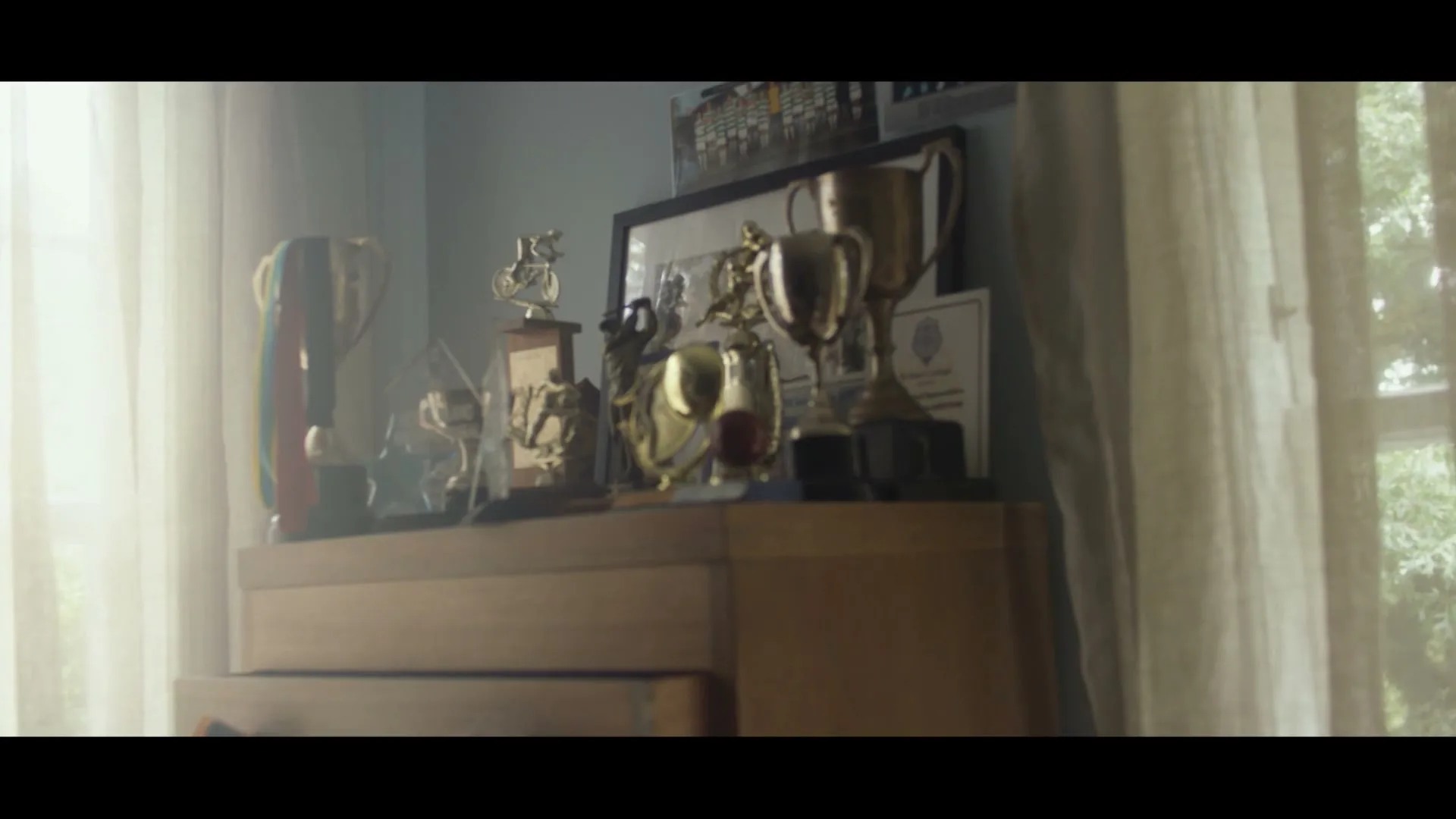 A collection of trophies and awards sits on a wooden dresser, illuminated by soft light from a nearby window, showcasing past achievements.