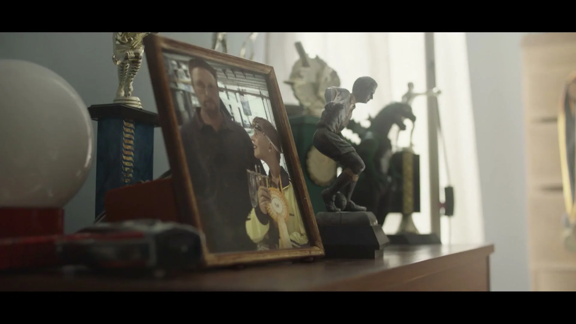 A framed photo of a man and a child holding a trophy sits on a wooden dresser next to several sports trophies in a softly lit room.
