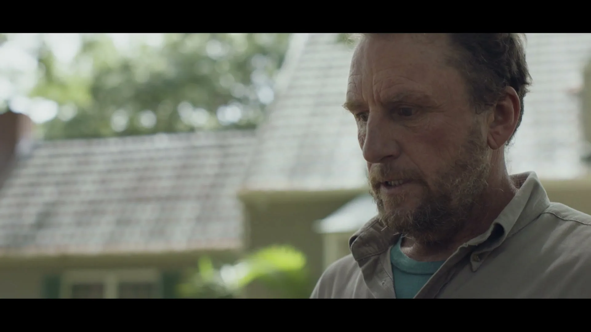A middle-aged man with a beard, wearing a collared shirt, looks down and then smiles, standing outdoors in front of a house.