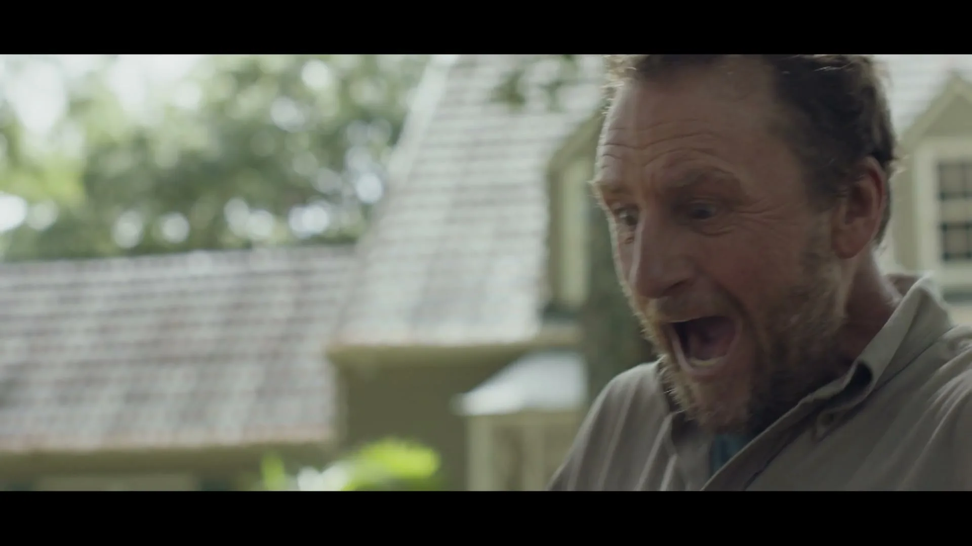 A middle-aged man with a beard, wearing a casual shirt, reacts with a surprised, open-mouthed expression in a suburban backyard.