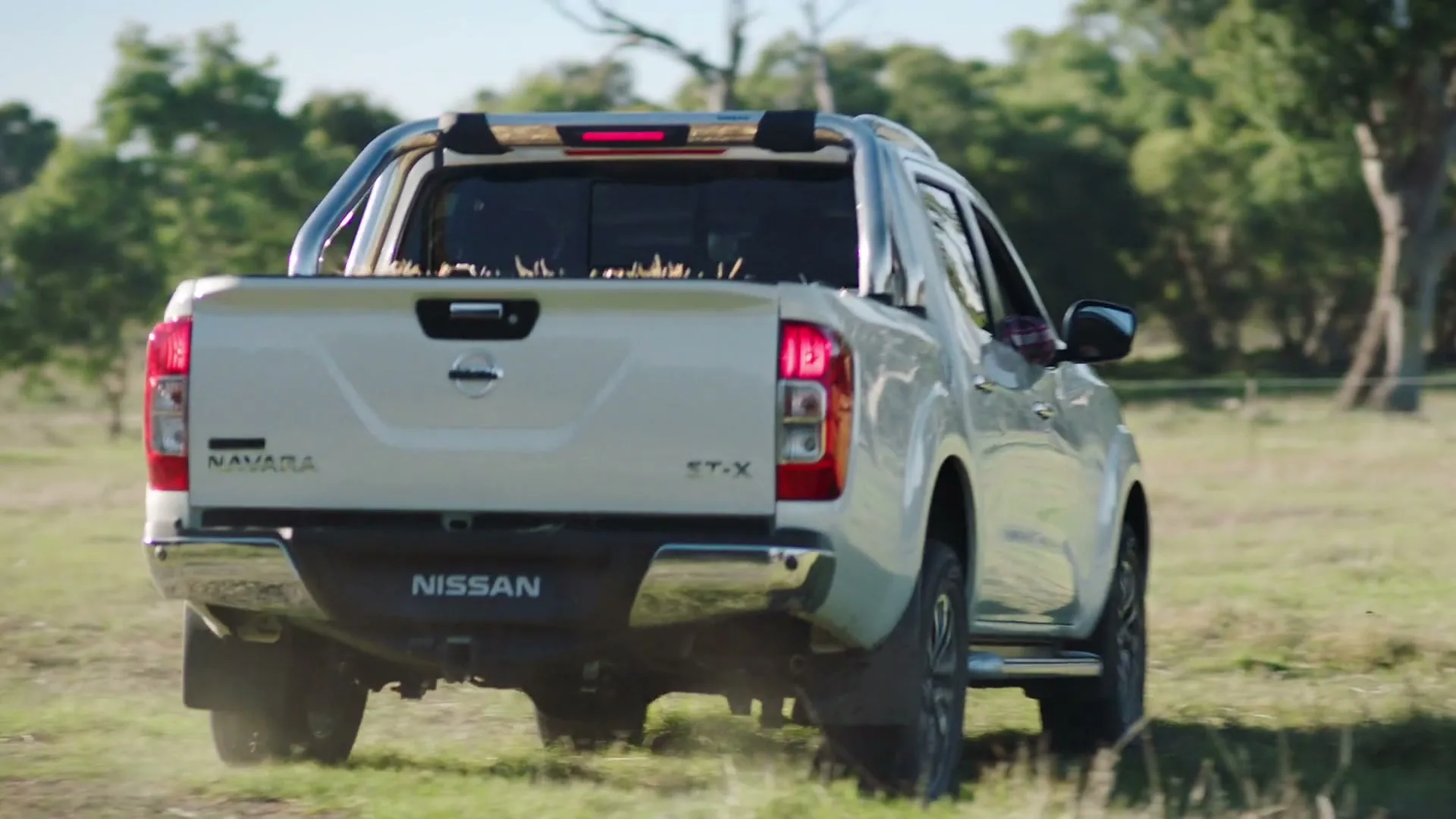 A white Nissan Navara pickup truck drives away from the camera through a grassy field with trees in the background on a sunny day.