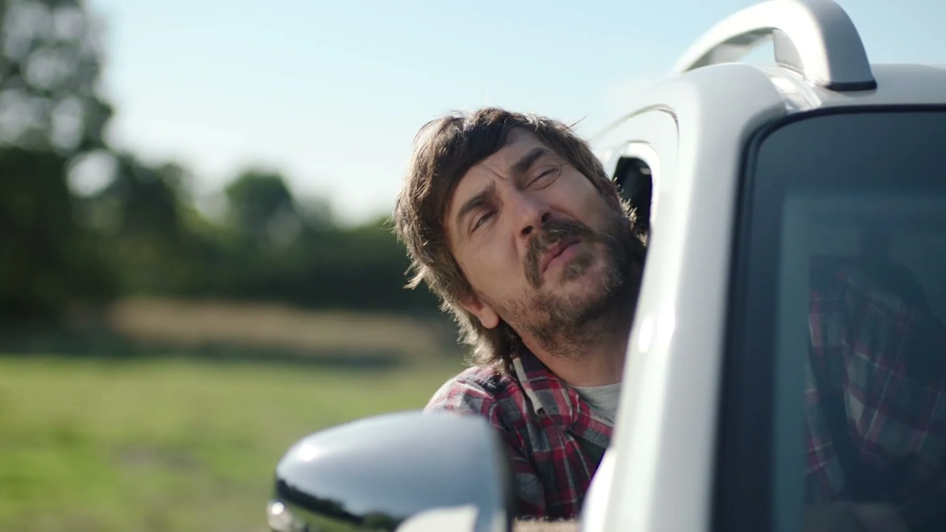 A man with a beard and plaid shirt looks out the window of a white truck, squinting in the sun in a rural setting.