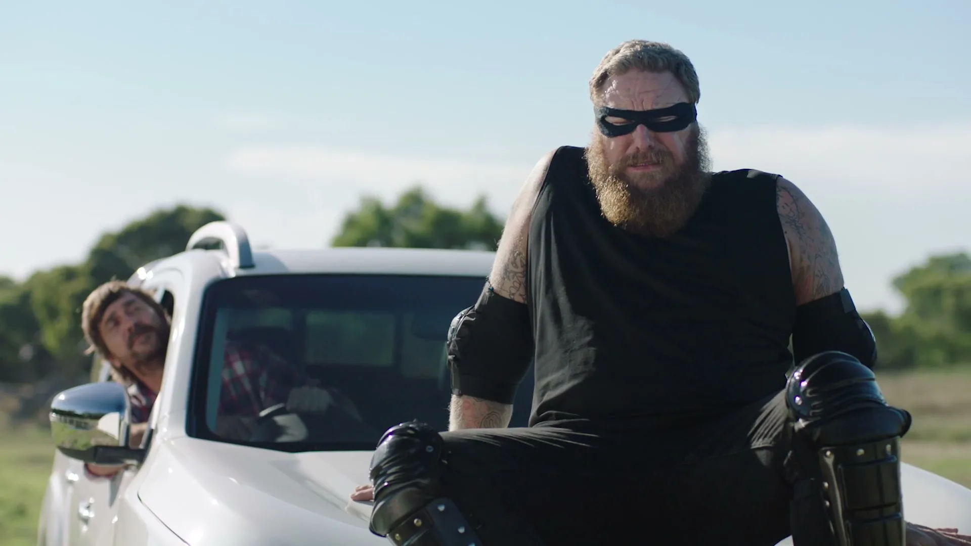 A man in a black superhero costume with a beard and mask sits on the hood of a white truck, while another man peeks out the driver's side window in a rural field.