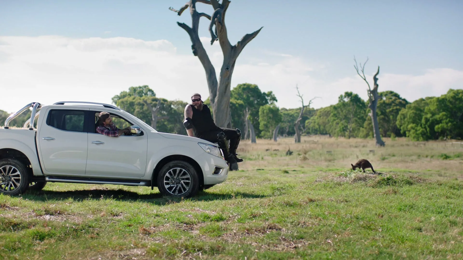 A man in a white pickup truck watches another man sitting on the hood, who is talking to a small kangaroo in a grassy field.