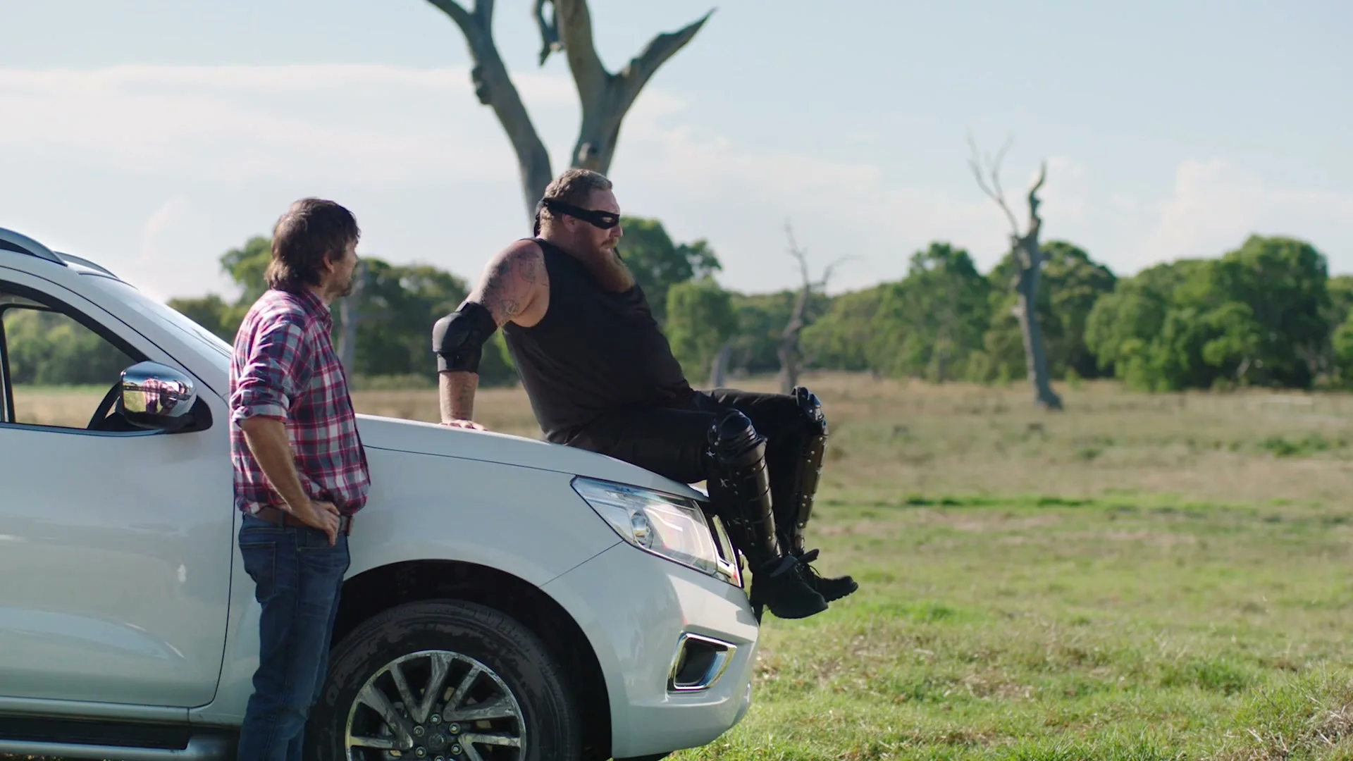 A man in a plaid shirt stands next to a white pickup truck, while another man in black clothing and sunglasses sits on the hood, in a grassy field with trees in the background.