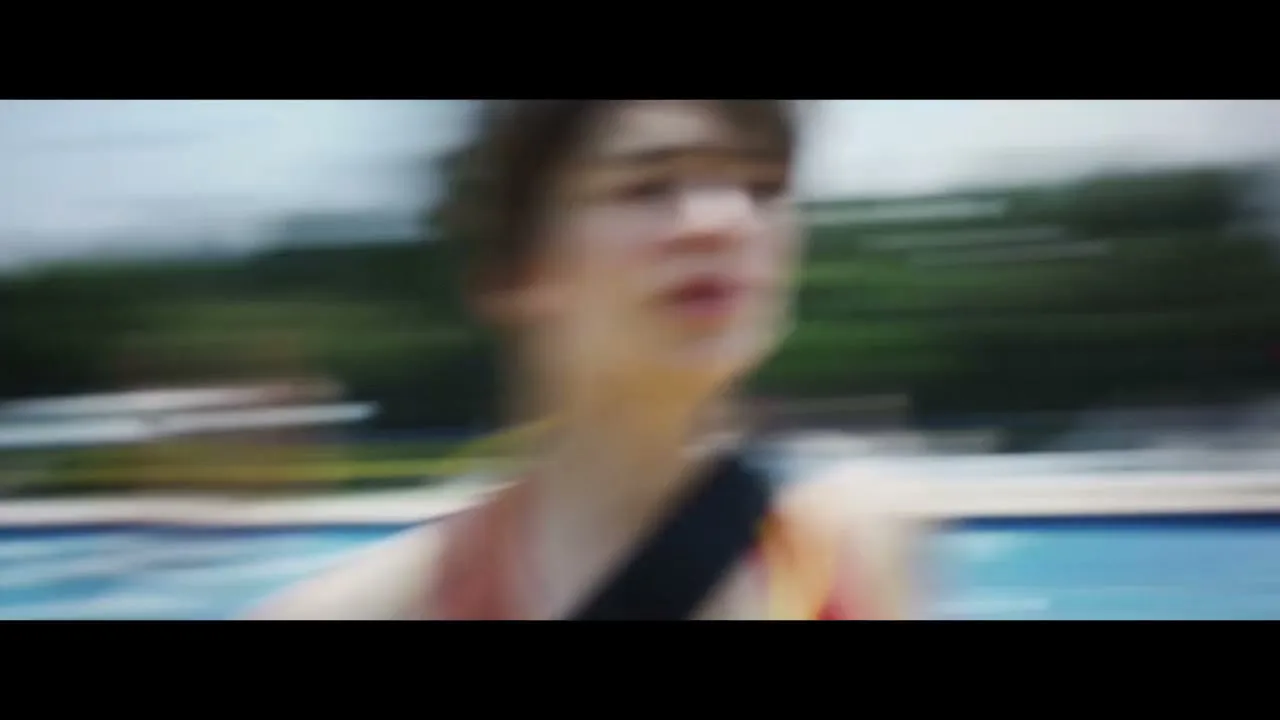 A female lifeguard in a red swimsuit runs towards the camera, holding a rescue buoy, then dives into a swimming pool on a sunny day.
