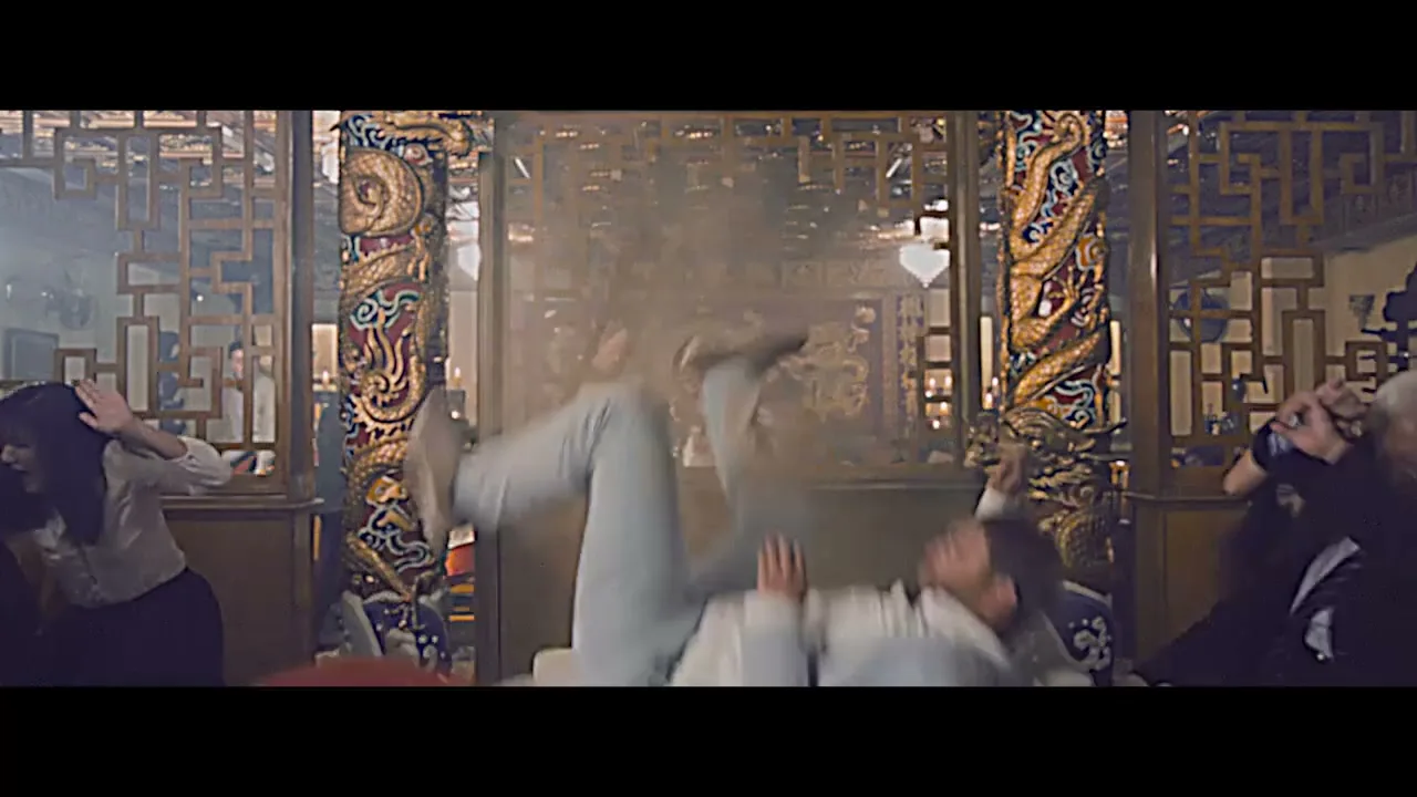 A man in a white martial arts uniform is thrown through a wooden screen in a busy Chinese restaurant, as other diners react in shock.