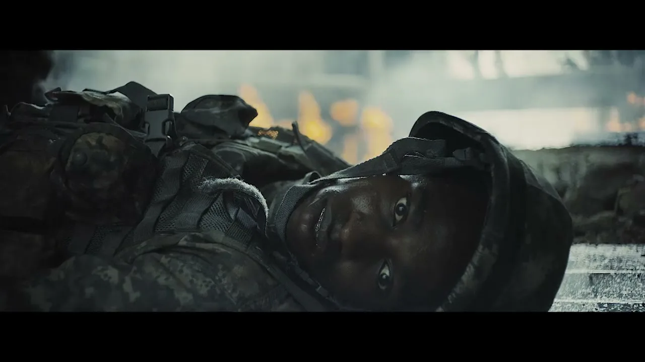 A young male soldier in camouflage uniform lies on the ground, looking up with a scared expression, with flames burning in the background.