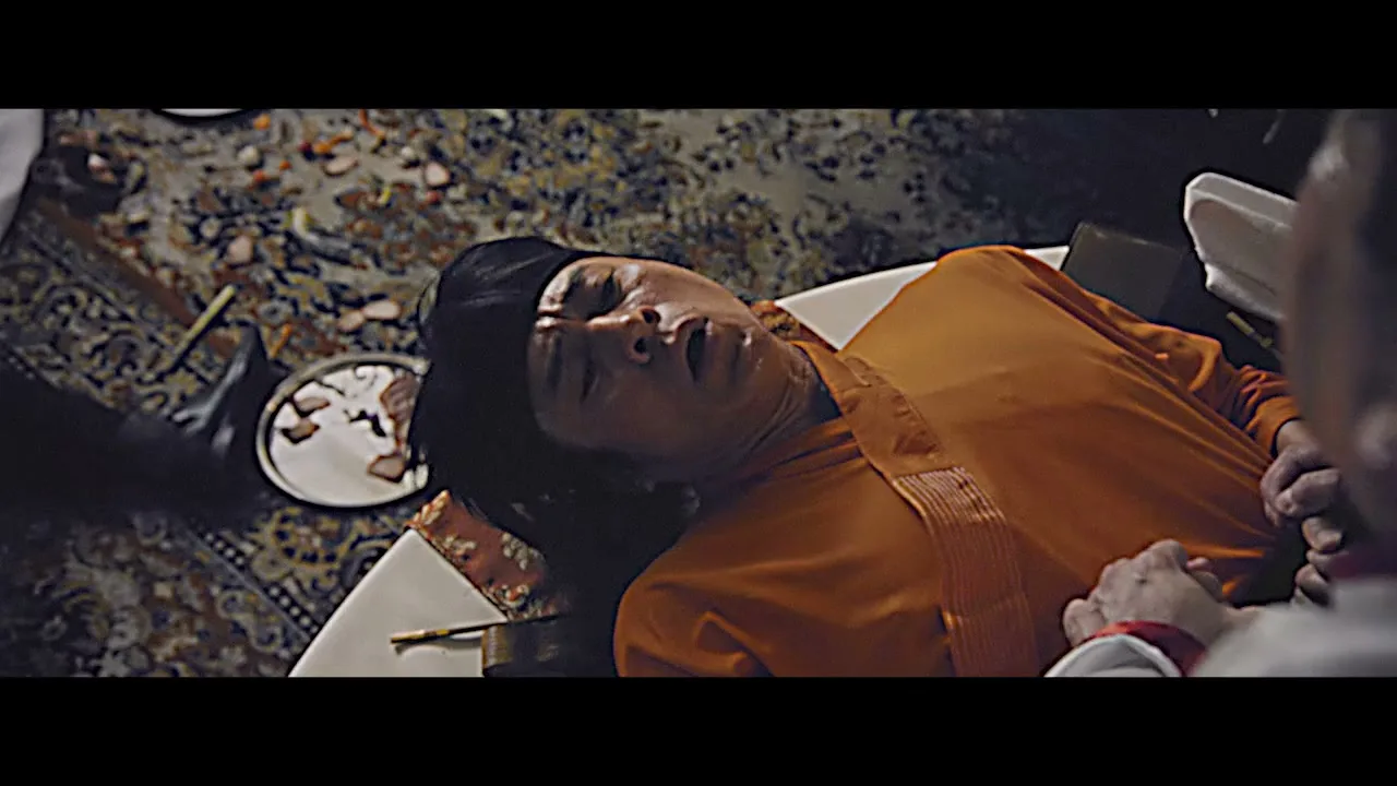 A man in an orange robe lies on a white surface, seemingly unconscious, while another person's arm is visible in the foreground, in a dimly lit interior.