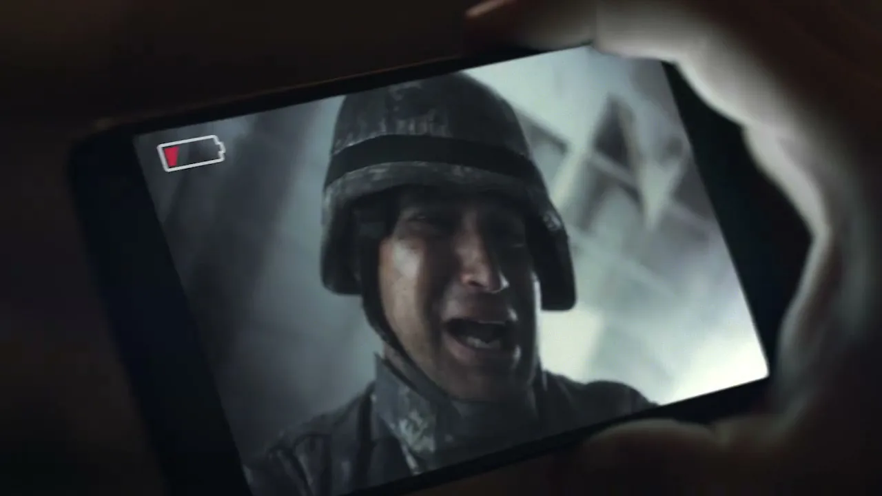 A close-up shot of a male soldier in a helmet and camouflage uniform, crying on a smartphone screen with a low battery icon, held by a hand in a dark setting.