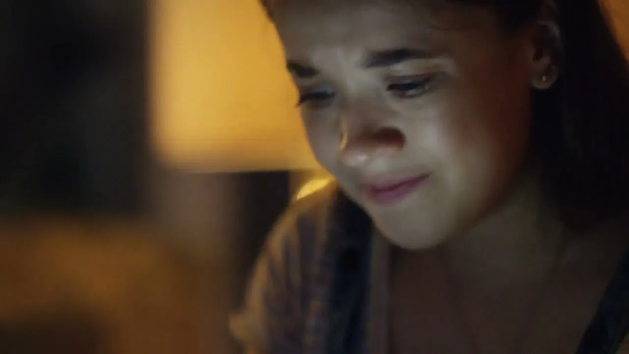 A young female with dark hair looks down with a worried expression, illuminated by a screen in a dark interior.