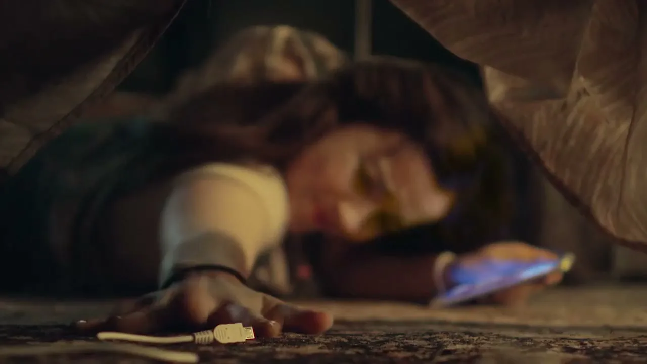 A young woman with long brown hair reaches for a white charging cable and a smartphone on a patterned carpet, under what appears to be a bed or blanket fort.