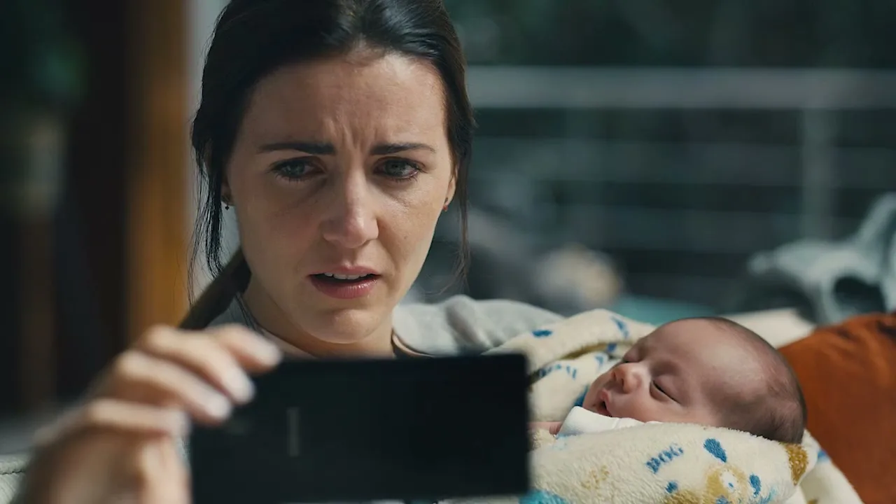 A worried mother holds her baby and a smartphone, looking distressed as she tries to use the device in an interior setting.