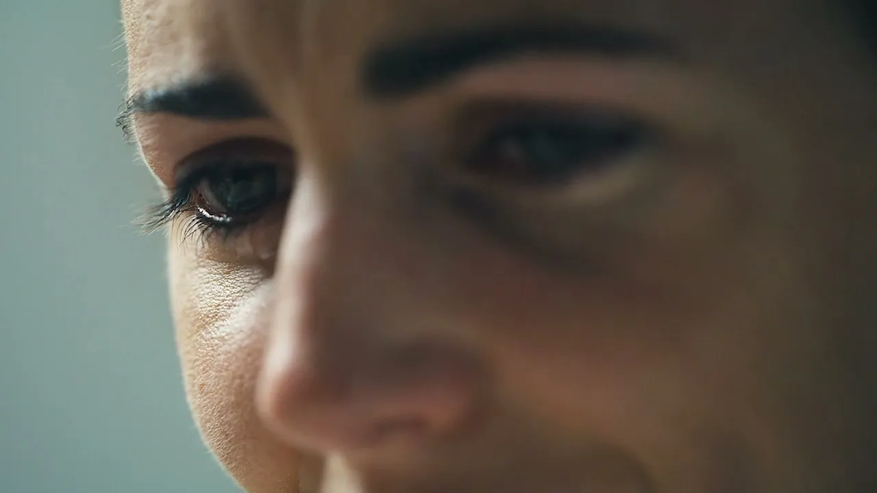 A woman with dark hair and a tear in her eye looks down, in a close-up shot.