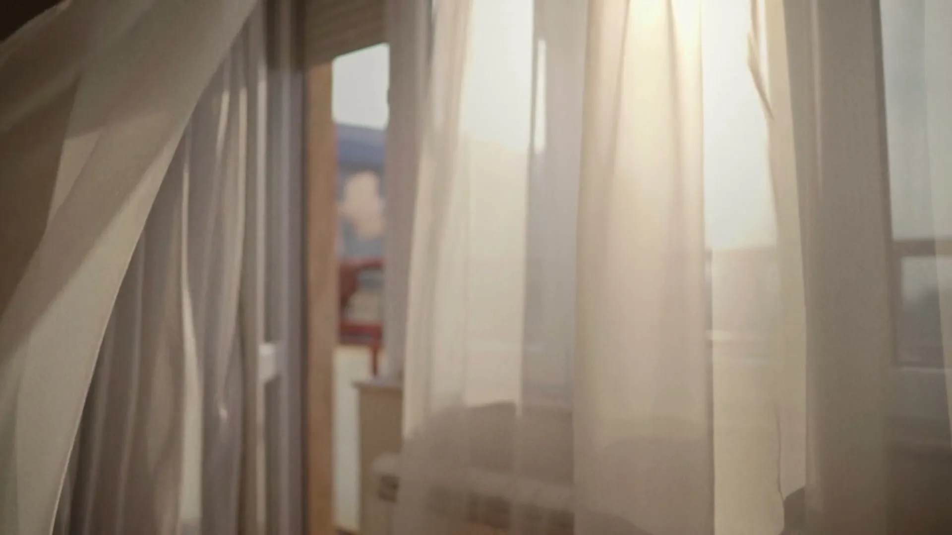 Sunlight streams through sheer white curtains blowing in the breeze from an open balcony door in a bright room.