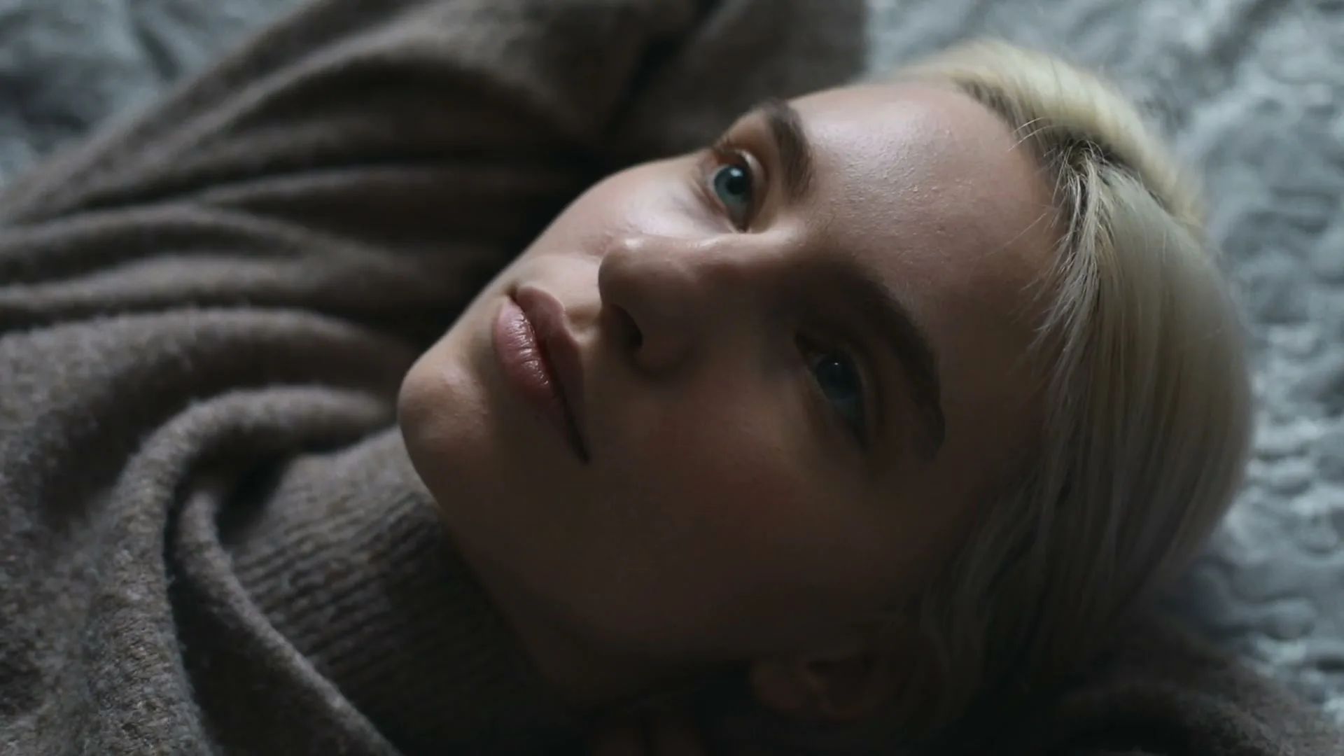 A young woman with blonde hair and blue eyes lies on a bed, looking up with a neutral expression in a medium closeup shot.