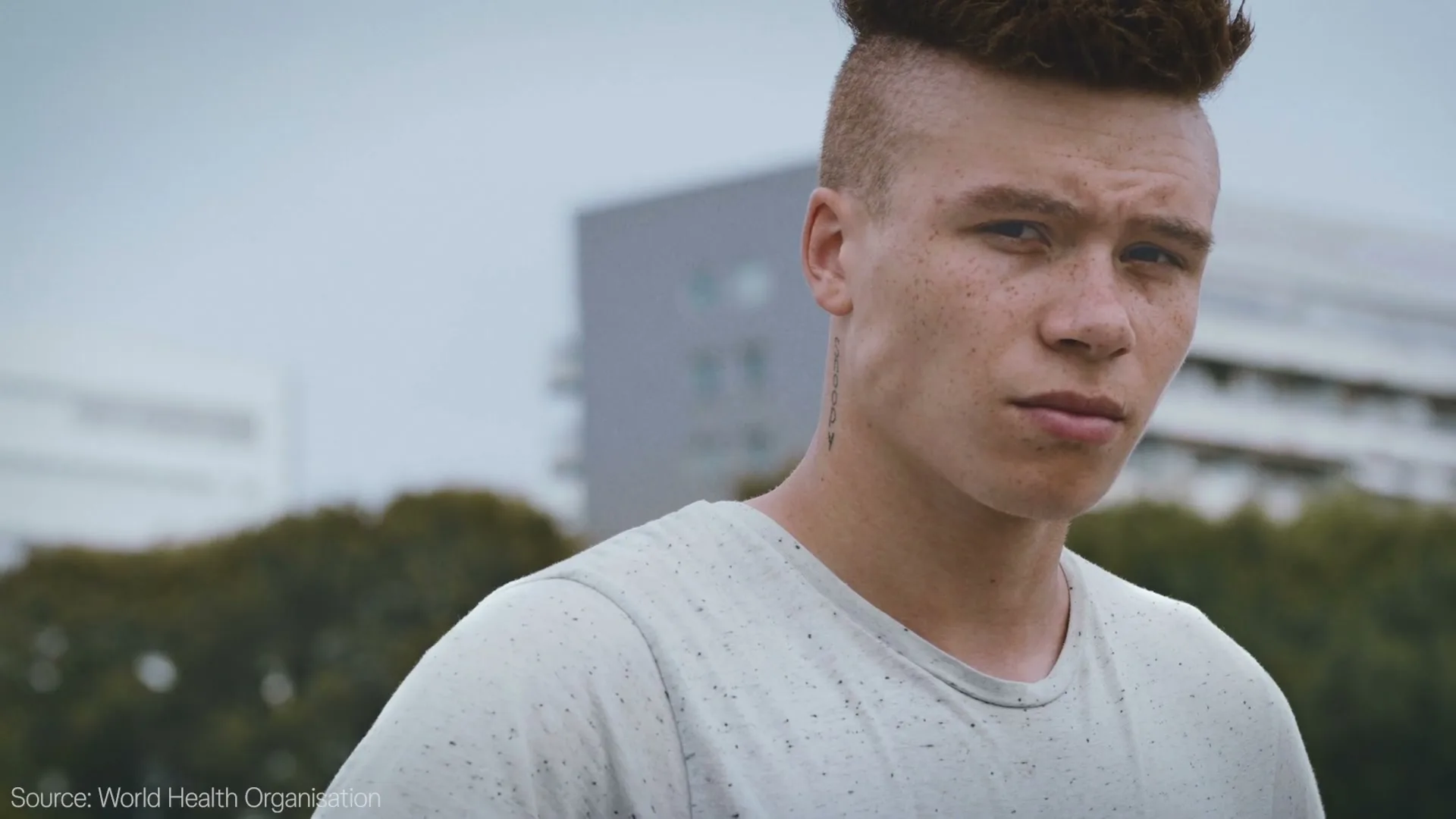 A young man with a fade haircut and freckles looks directly at the camera with a serious expression, outdoors with buildings in the background.